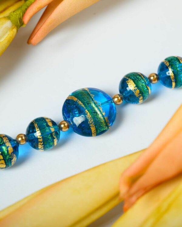 Close-up of a Murano necklace with vibrant blue and green beads featuring gold accents, displayed on a decorative surface